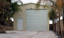 A tan-colored storage shed features a white door at the end of a driveway. The metal shed extends the garage with a frame made of bent pipe.